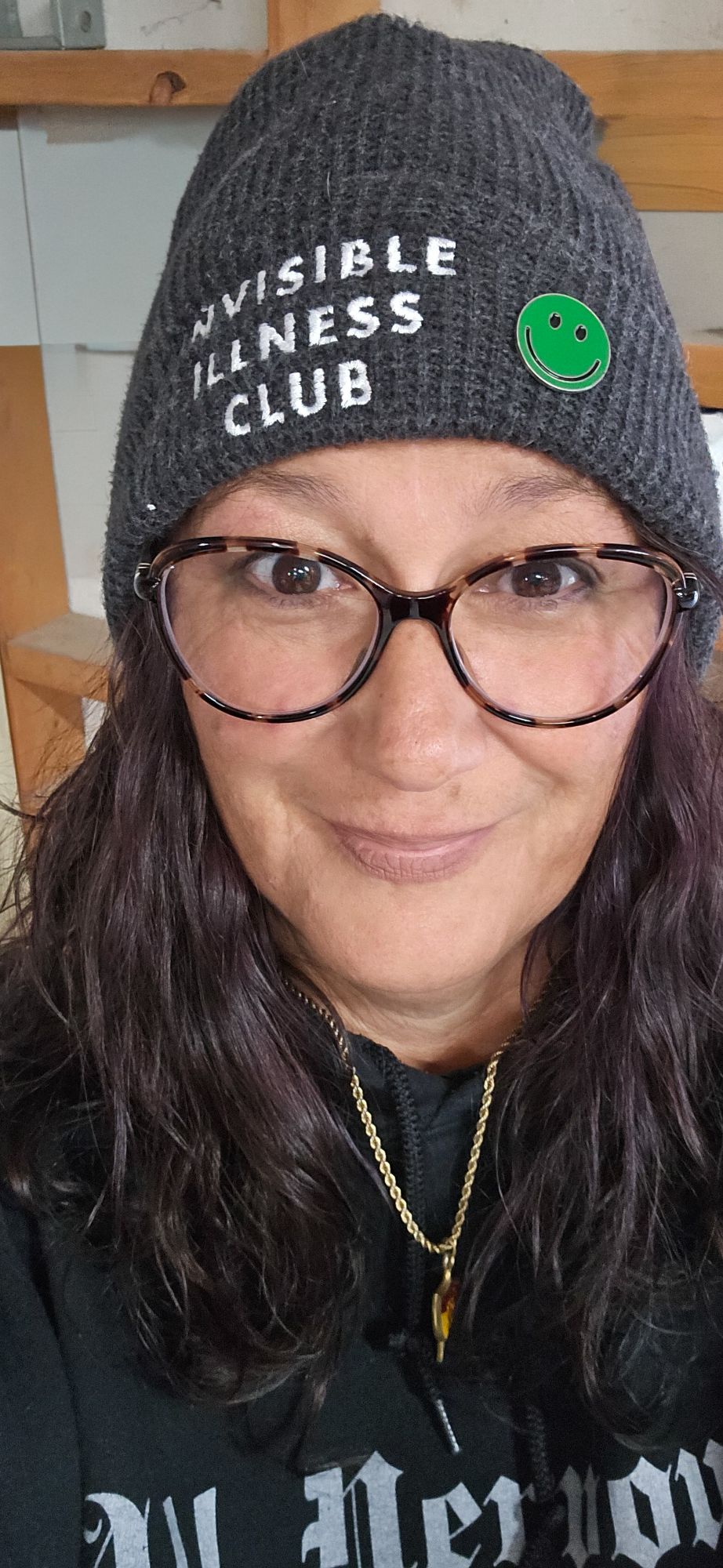 Momma Rich smiles at the camera. She is wearing a gray stocking cap with "Invisible Illness Club" embroidered in white and a green smiley face pin that represents Mental Health Awareness. She is wearing brow tortoise shell glasses and a black sweatshirt that says, "All Nervous, No System" and her husband's senior key.